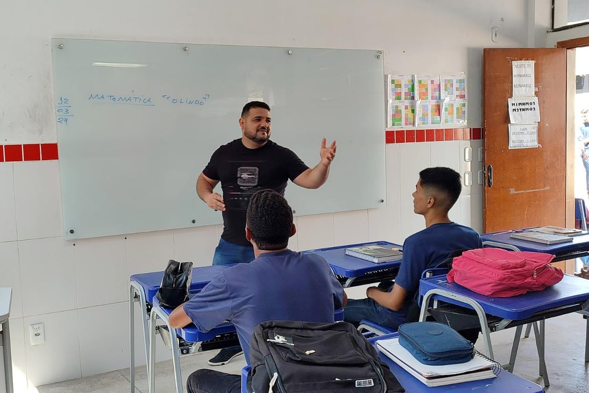 Professor em sala de aula, com estudantes atentos e quadro ao fundo