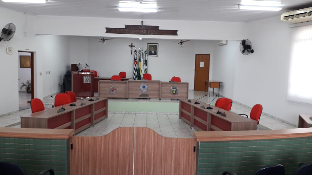 Interior do plenário da Câmara Municipal de Santa Adélia, com mesas e cadeiras em disposição para sessão, em ambiente administrativo público