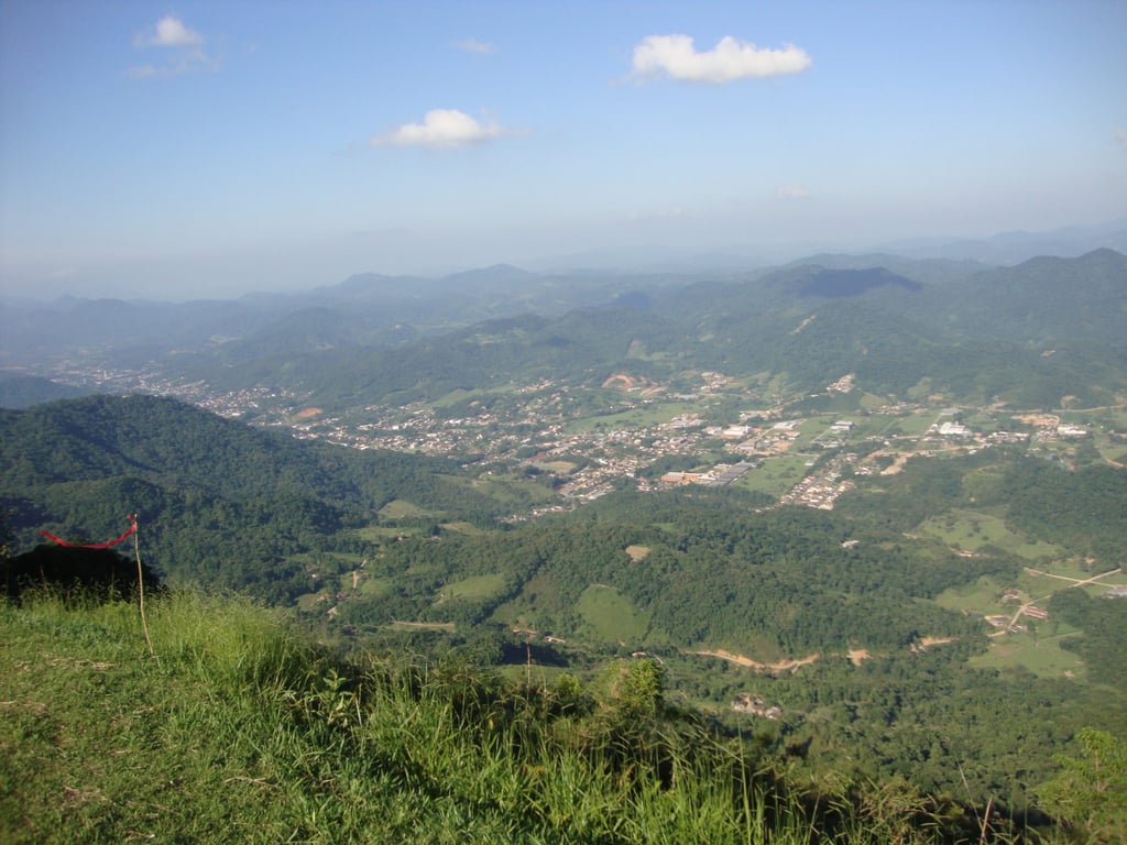 Vista aérea do Vale do Itajaí, em Santa Catarina, com montanhas e vegetação. A cena mostra uma paisagem natural com áreas urbanizadas ao fundo.