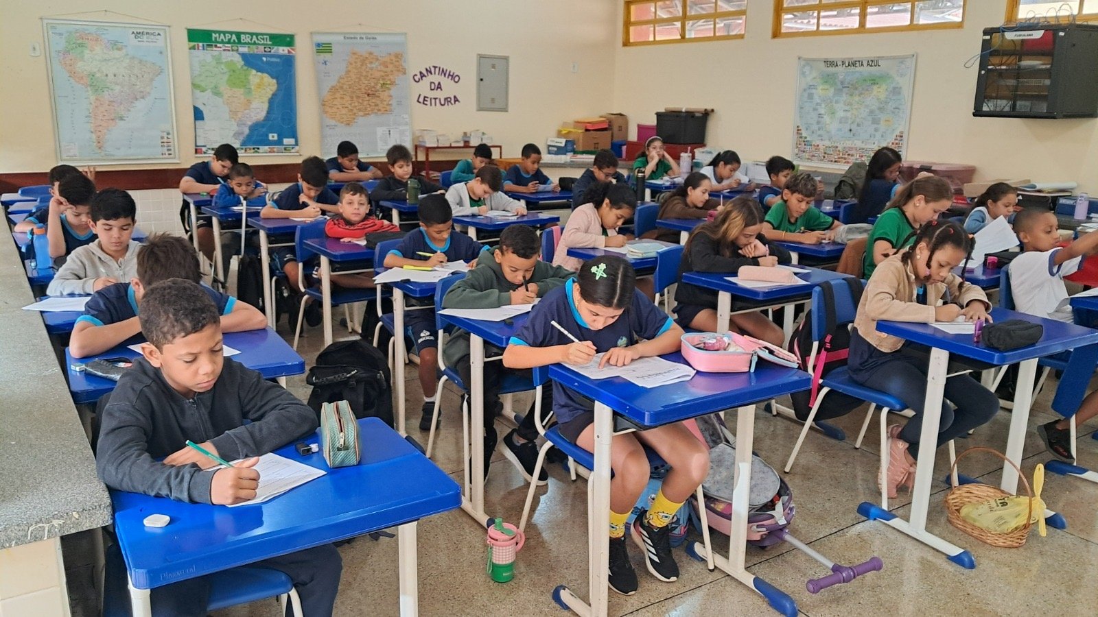 Sala de aula pública com alunos, carteiras organizadas e materiais pedagógicos; ambiente iluminado por luz natural