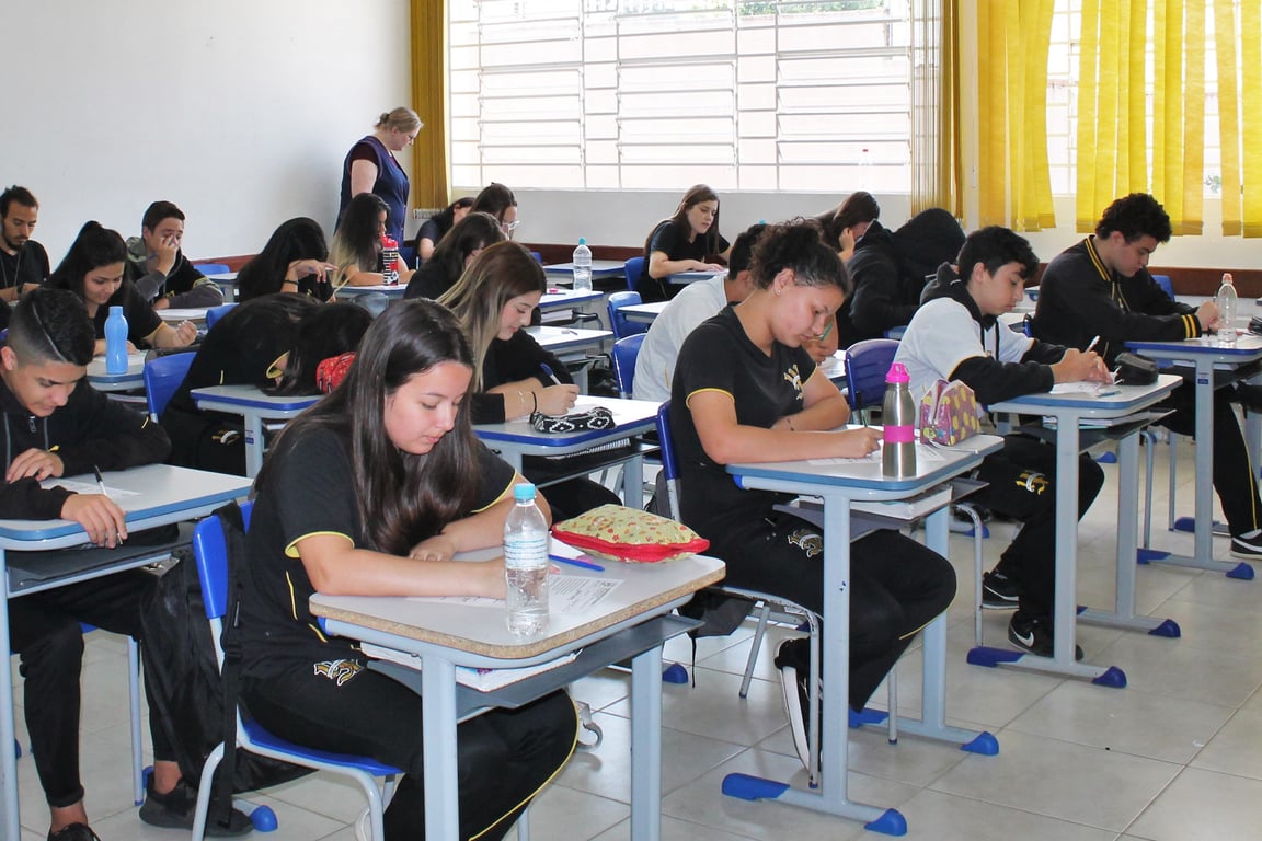 Sala de aula da rede pública com estudantes em prova, carteiras alinhadas e quadro ao fundo