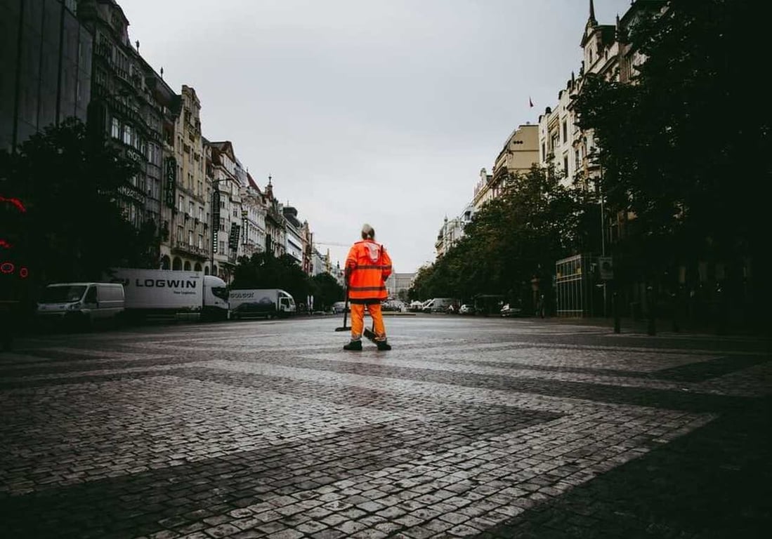 Equipe de limpeza urbana varrendo rua com coletes e EPIs em uma cidade. A cena apresenta um ambiente urbano arborizado.