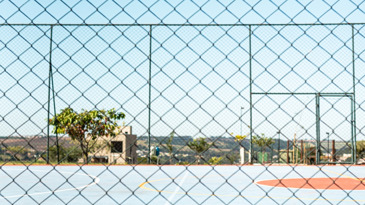 Quadra poliesportiva coberta vazia, com marcações de futsal e basquete e arquibancada simples