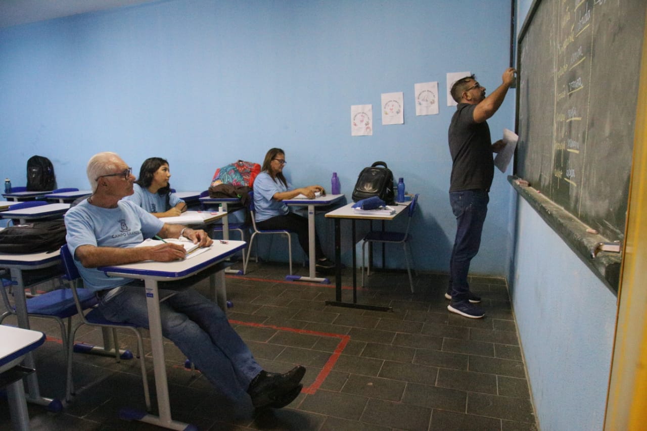 Sala de aula de EJA com adultos estudando, quadro e materiais didáticos ao fundo