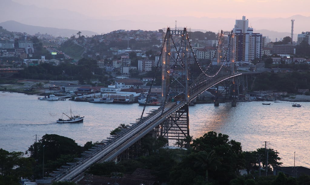 Vista aérea da ponte Hercílio Luz ao entardecer, com skyline e mar de Florianópolis ao fundo