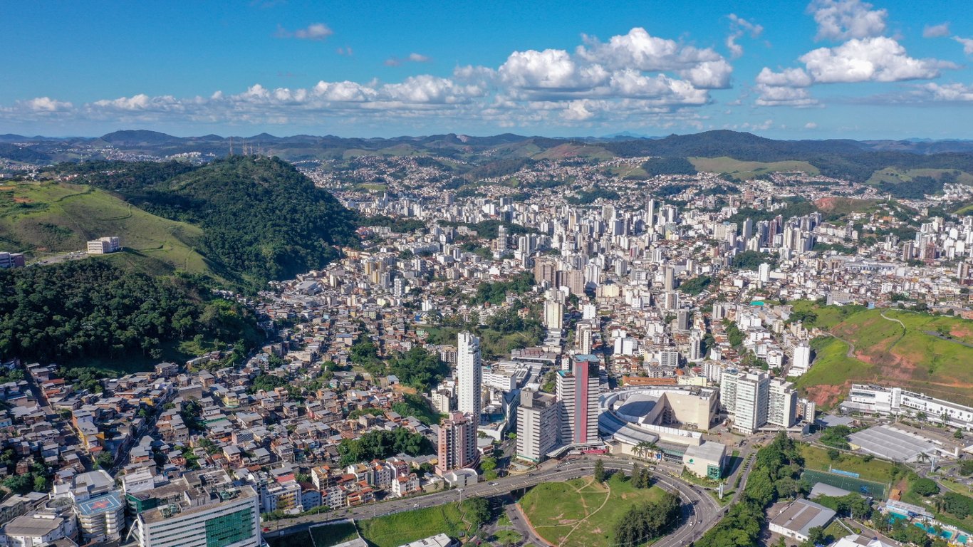 Vista aérea de Juiz de Fora MG, com prédios e morros verdes ao fundo, em dia de céu claro