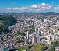 Vista aérea de Juiz de Fora MG, com prédios e morros verdes ao fundo, em dia de céu claro
