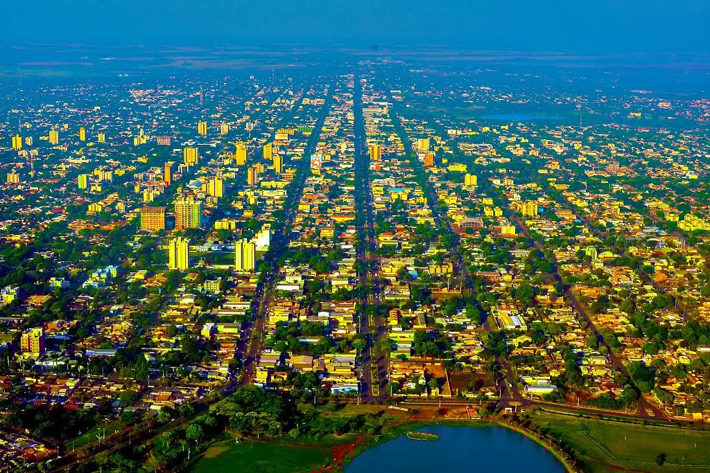 Vista aérea de uma cidade do interior de MS ao entardecer, com ruas arborizadas e construções baixas.