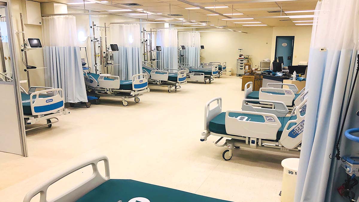 Corredor de hospital com várias camas e cortinas; ambiente profissional e organizado.