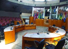 Interior de plenário de câmara municipal de pequeno porte, com cadeiras e bandeiras em disposição típica de sessões legislativas.