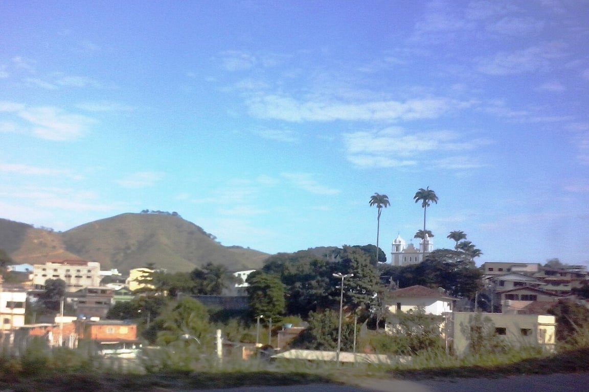 Vista urbana em vale cercado por morros verdes, céu azul; atmosfera do interior capixaba