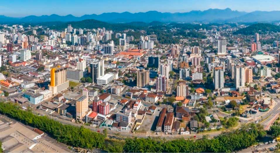 Vista aérea de Joinville, com área urbana e morros ao fundo, composição horizontal