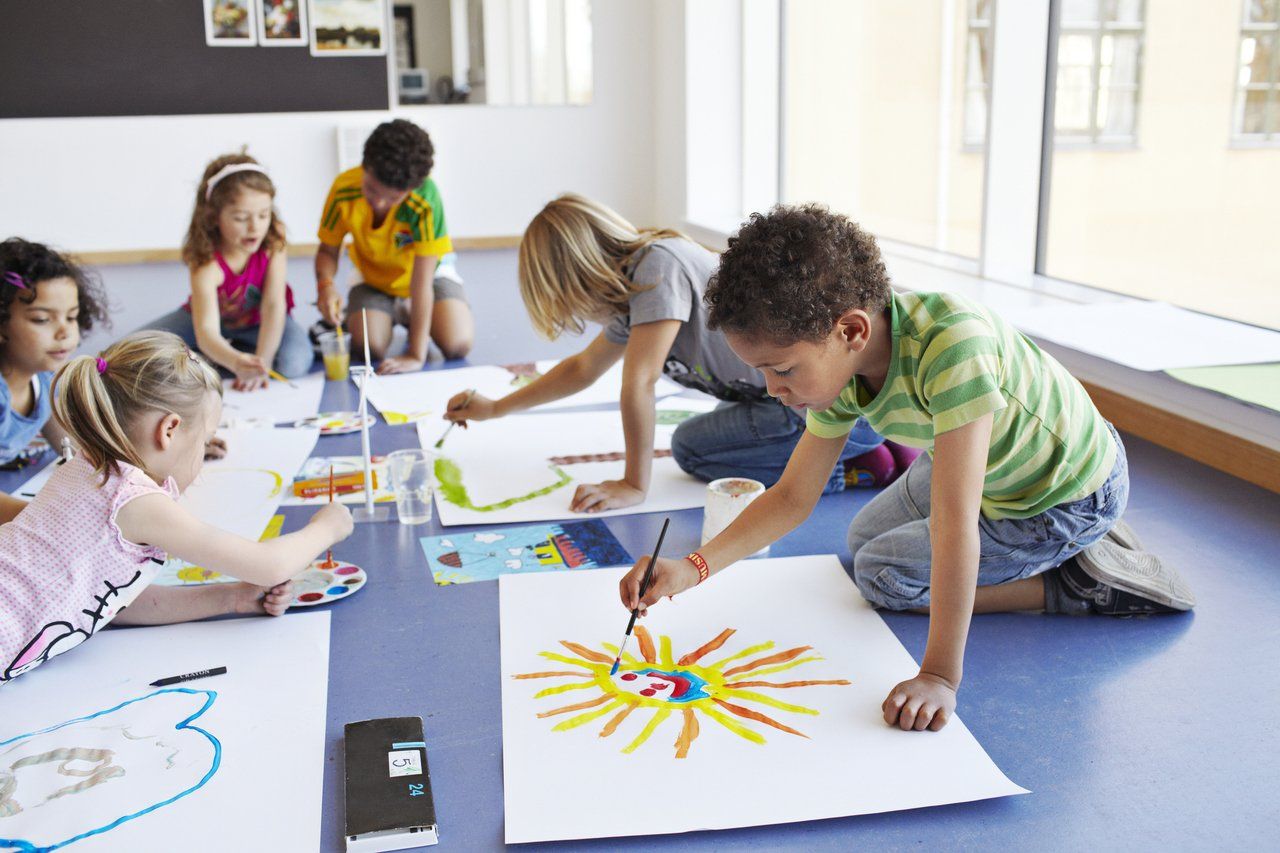 Sala de aula de Educação Infantil com crianças pintando e professora auxiliando, em ambiente iluminado e acolhedor.