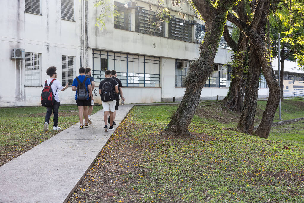 Estudantes em ambiente de campus universitário, prédios ao fundo, dia ensolarado