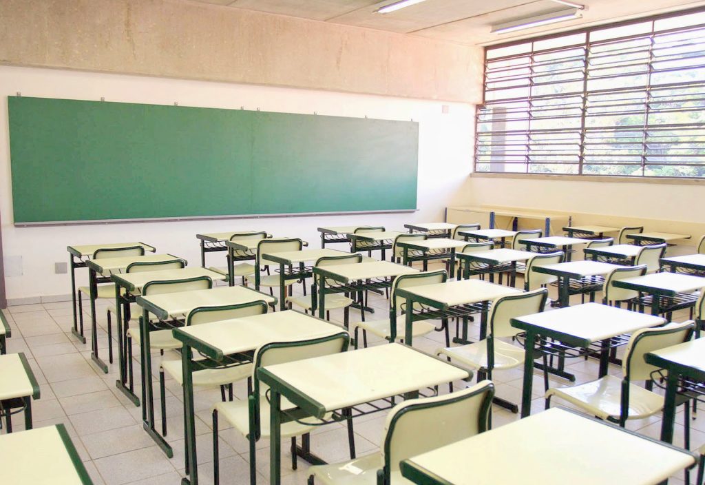 Sala de aula de escola pública com carteiras e quadro branco, sem alunos identificáveis