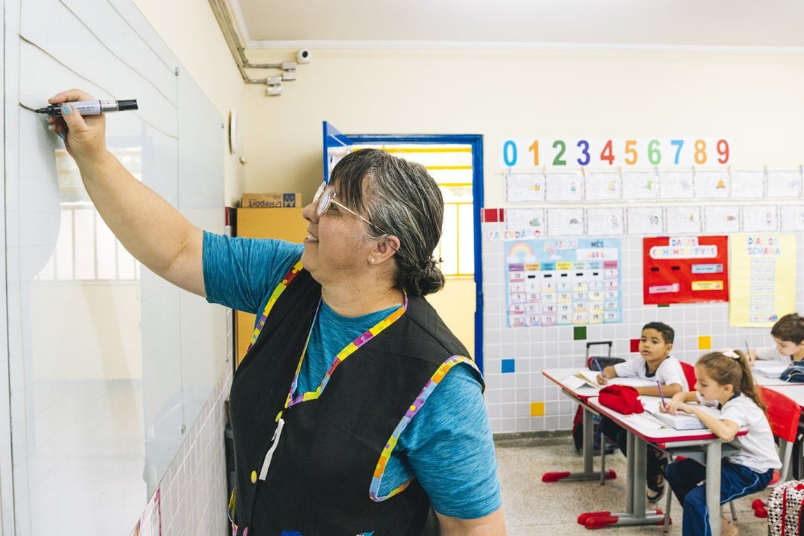 Sala de aula com professora escrevendo na lousa e estudantes ao fundo