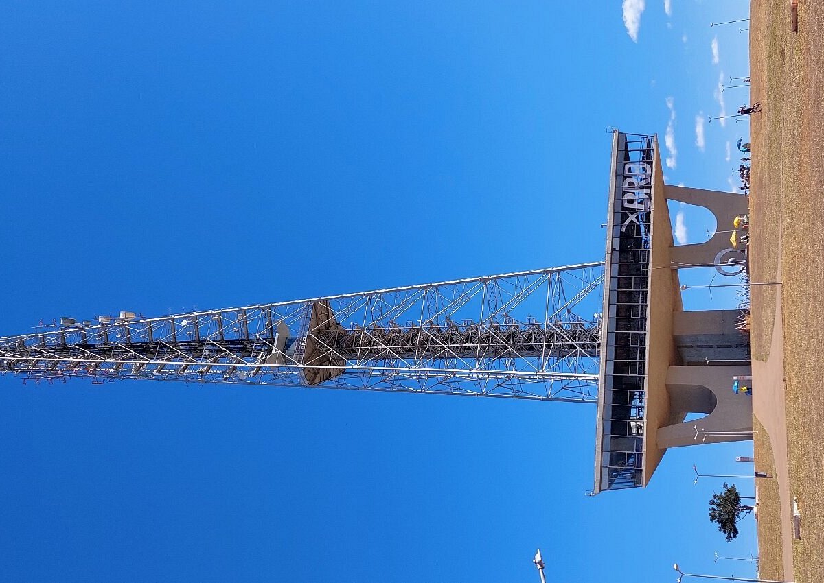 Foto do mirante da Torre de TV de Brasília, com céu azul e pessoas ao fundo, em um dia ensolarado.