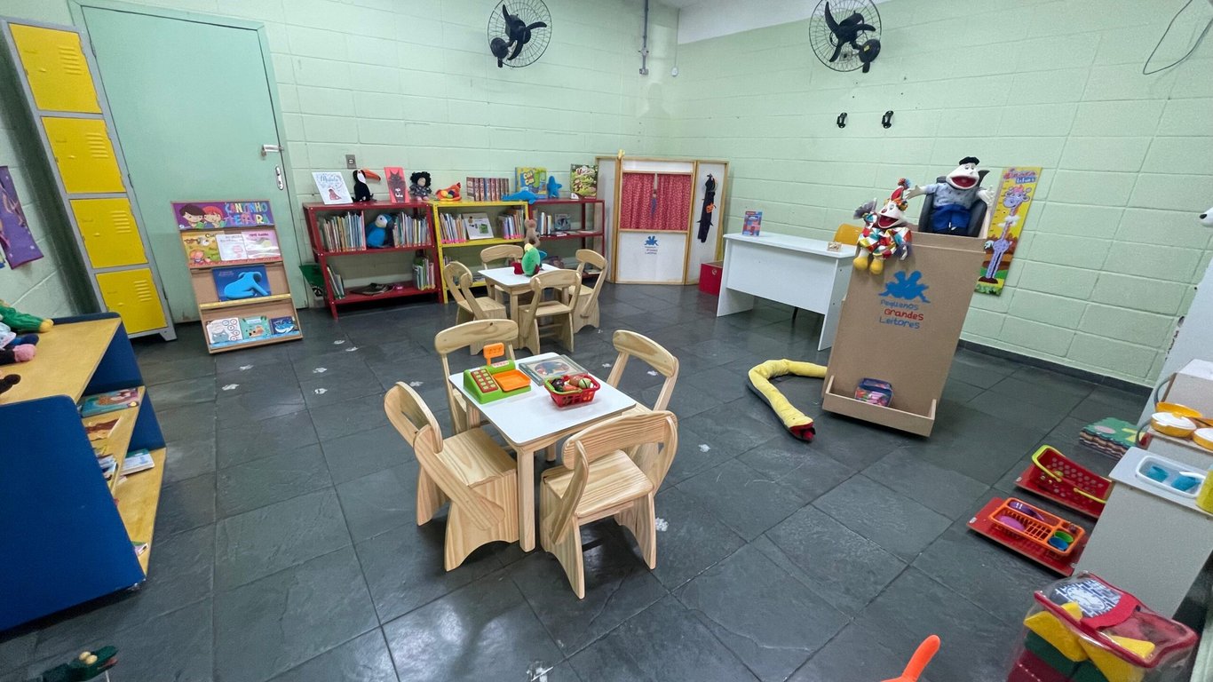 Interior de uma biblioteca escolar pequena, com estantes, mesas e cadeiras, ambiente iluminado e organizado, sem pessoas visíveis