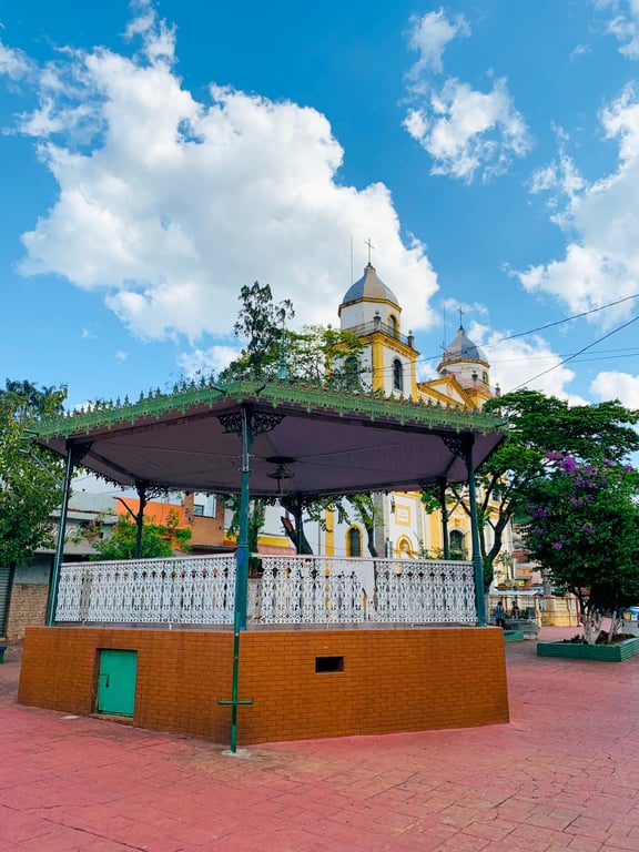 Coreto em praça arborizada de cidade do interior paulista, com igreja ao fundo