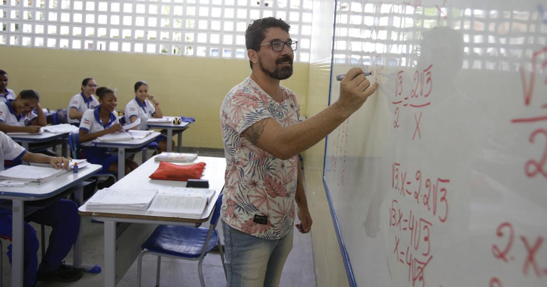 Professor escrevendo equações em um quadro branco em sala de aula, alunos ao fundo atentos.