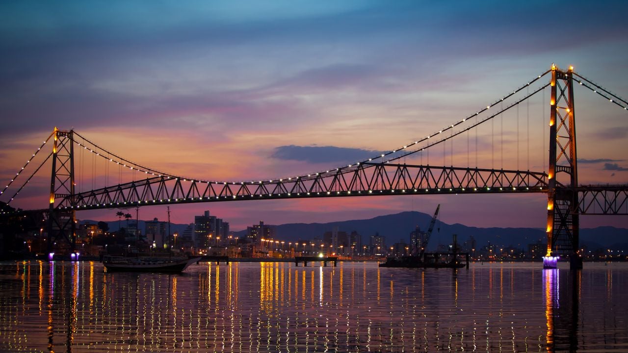 Pôr do sol na baía de Florianópolis, com skyline moderno e ponte Hercílio Luz ao fundo