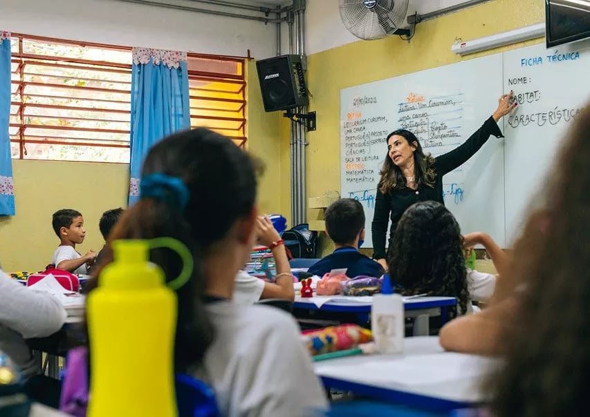 Sala de aula em escola pública, com professora no quadro e alunos em carteiras, em ambiente iluminado