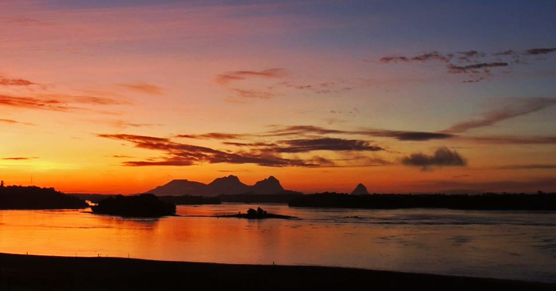 Pôr do sol refletido no Rio Negro, com vegetação amazônica e cadeias de montanhas ao fundo.