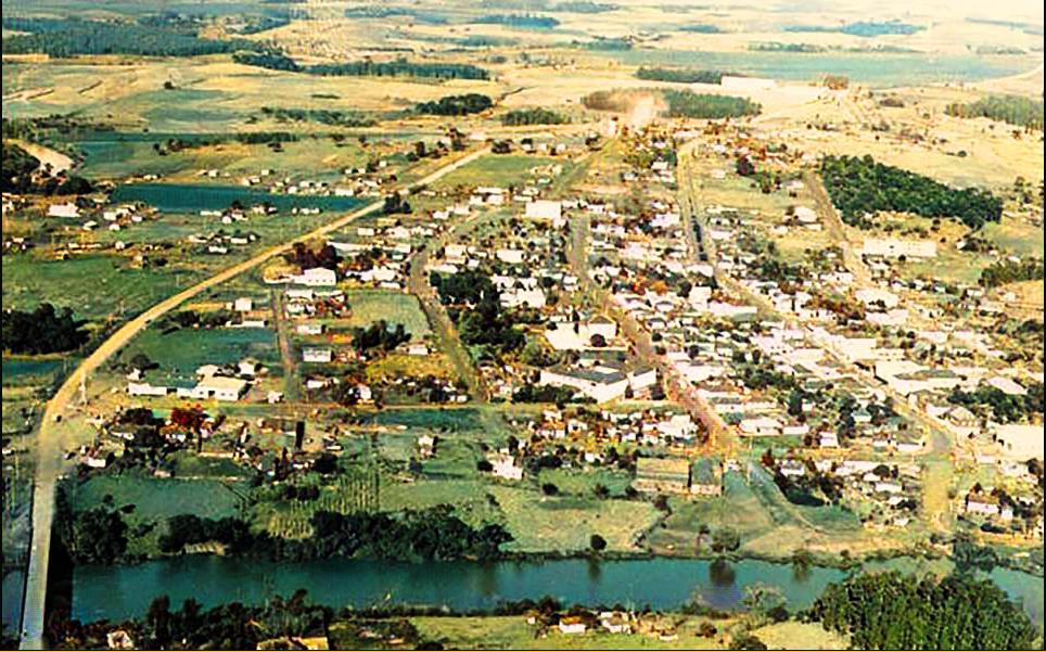 Vista aérea de cidade do interior do RS, com praça central e igreja, rodeada por área urbana e campos agrícolas
