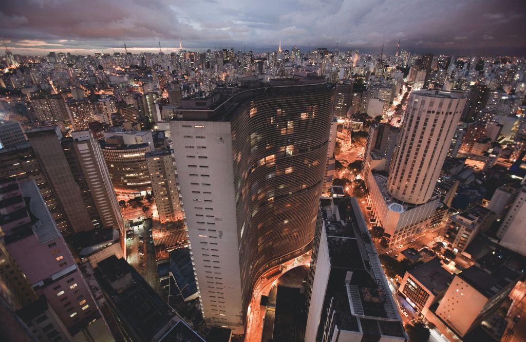 Vista panorâmica de São Paulo ao entardecer, com prédios iluminados e uma avenida movimentada. A imagem retrata a cidade em um momento vibrante e dinâmico.