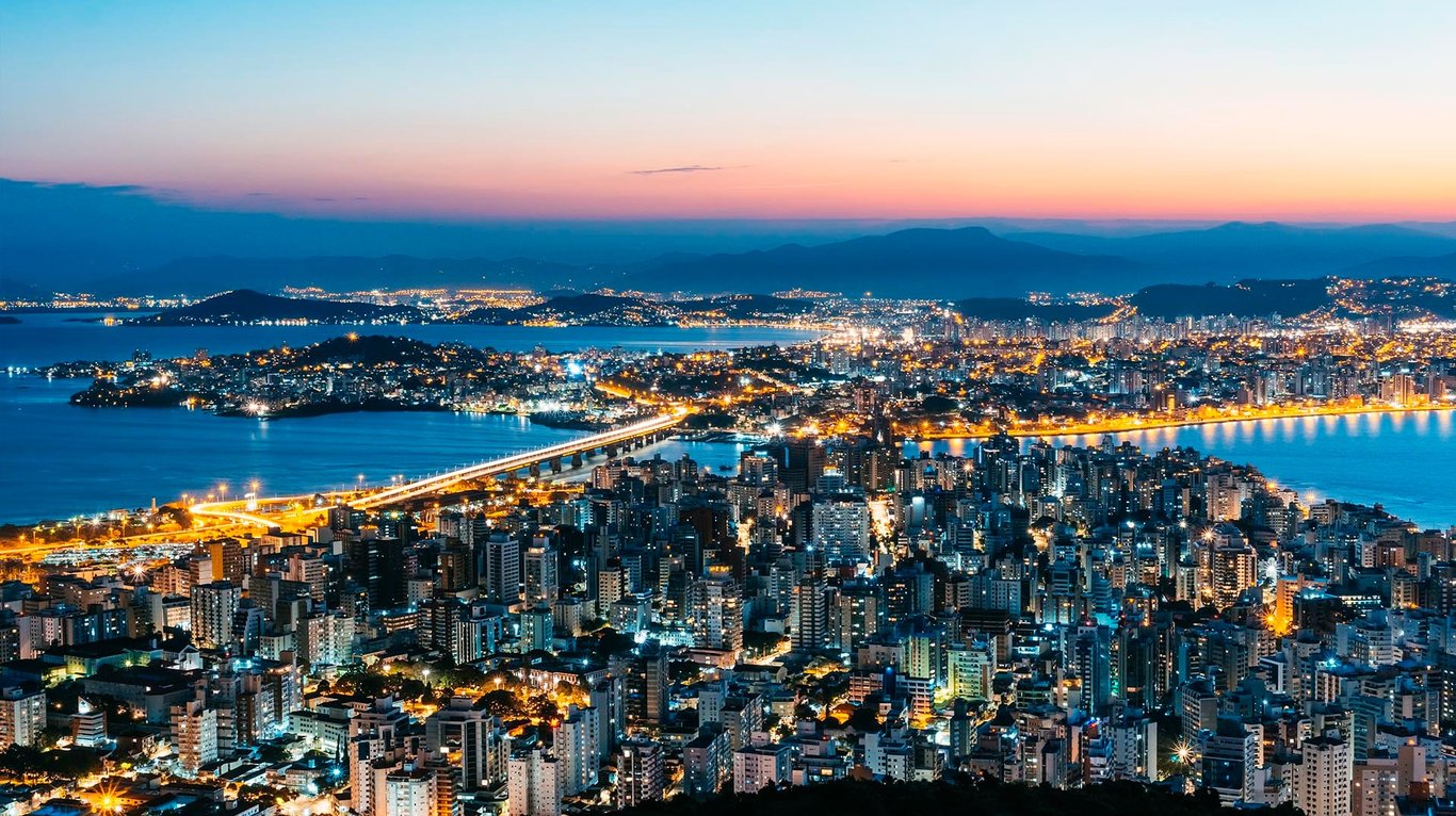 Vista aérea de Florianópolis ao entardecer, com pontes e orla iluminadas