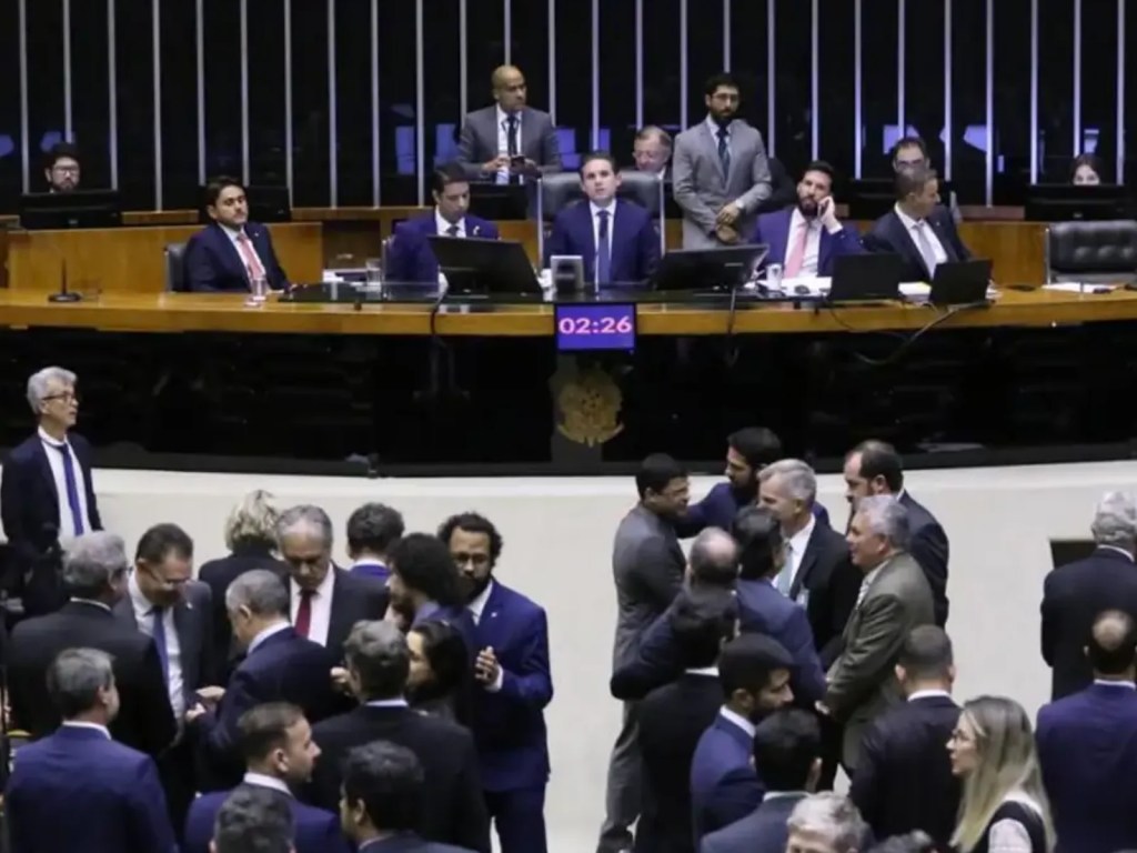 Ambiente interno de uma câmara legislativa no Brasil, com assentos e microfones, iluminação quente