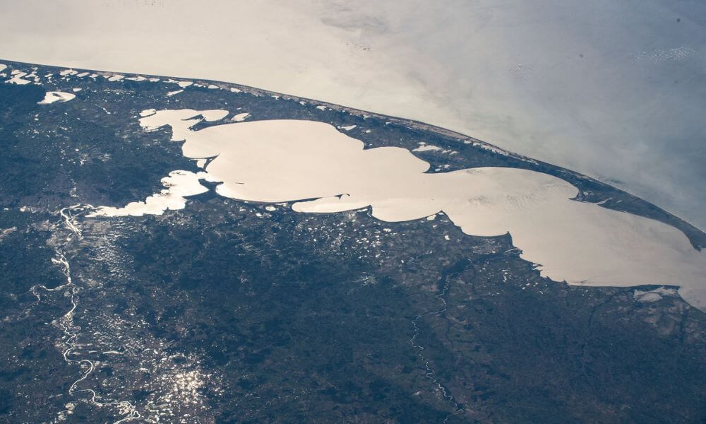Lagoa dos Patos na Costa Doce RS, em dia claro, com vegetação e orla ao fundo.