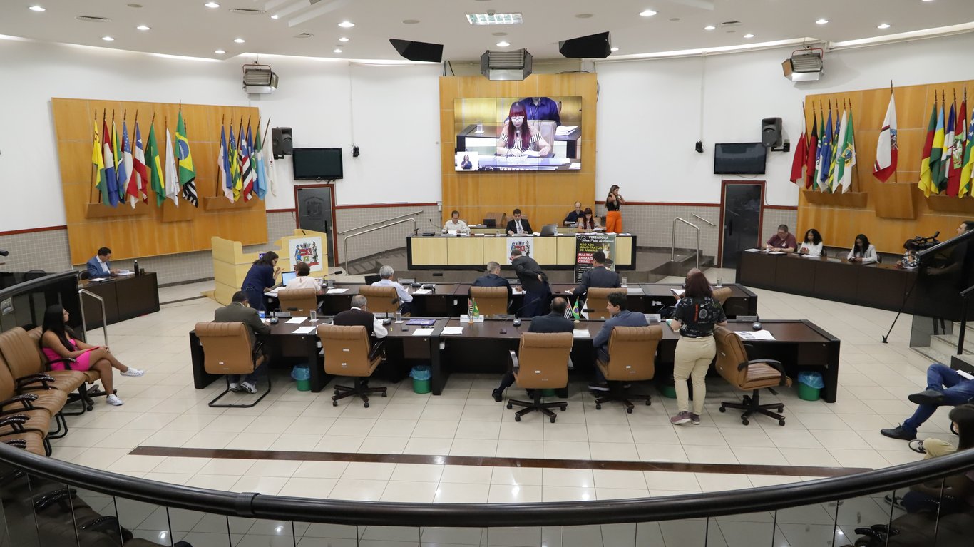 Interior de plenário de câmara municipal, com mesas, cadeiras e bandeiras ao fundo