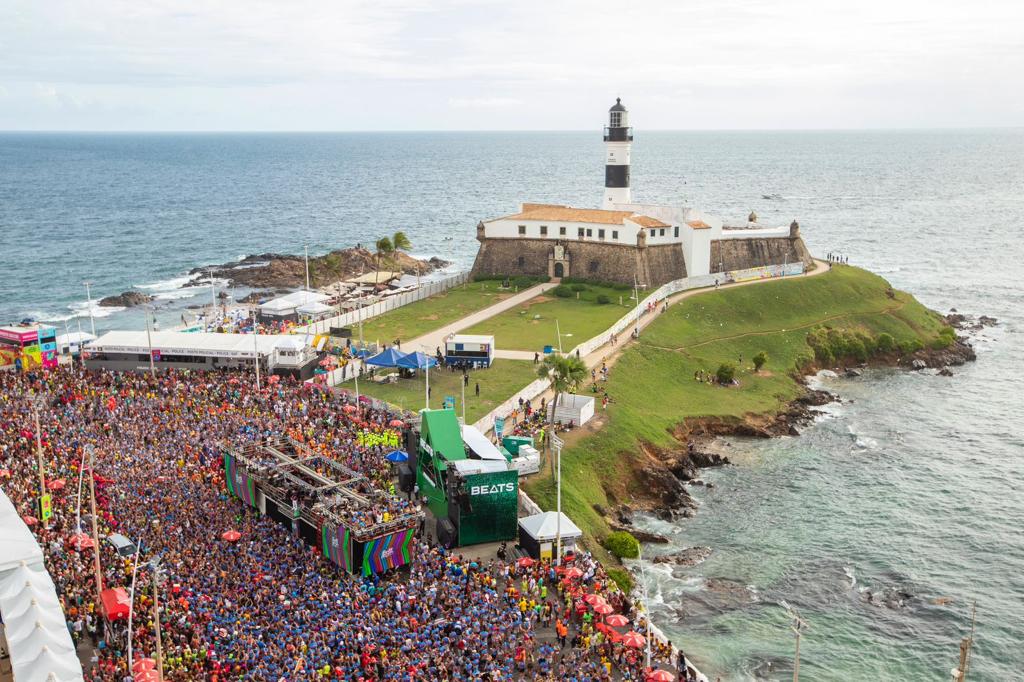 Vista aérea do circuito Barra-Ondina durante o Carnaval de Salvador, com trios elétricos e multidão ao longo da orla ao entardecer