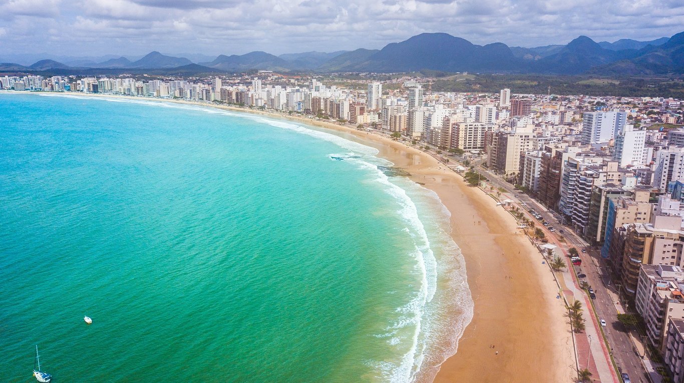 Vista aérea de praia em Guarapari, com mar azul-turquesa, longa faixa de areia e prédios ao fundo