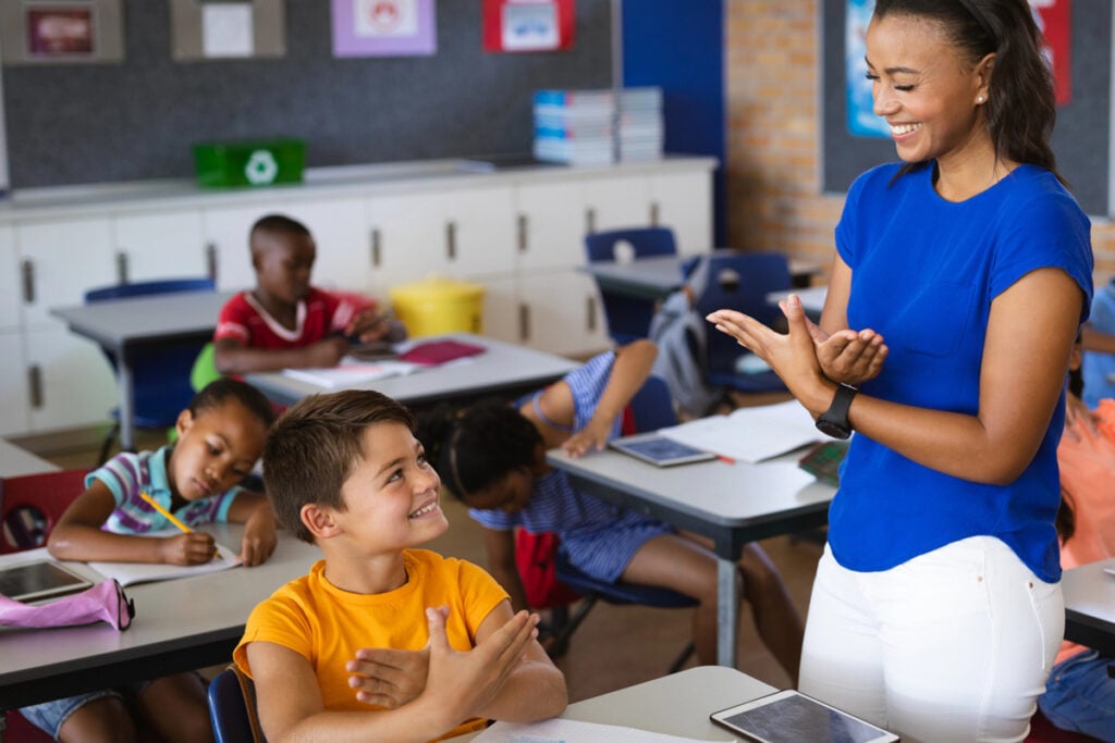 Sala de aula com professora realizando sinais de Libras; estudantes acompanham atentamente a demonstração.
