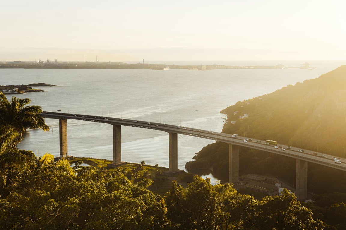 Vista aérea da Terceira Ponte em Vitória ES ao pôr do sol, com a baía ao fundo e vegetação nas laterais.