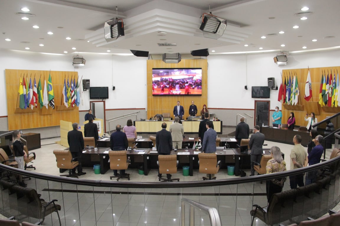 Plenário de uma câmara municipal, com vereadores em sessão e bandeiras ao fundo