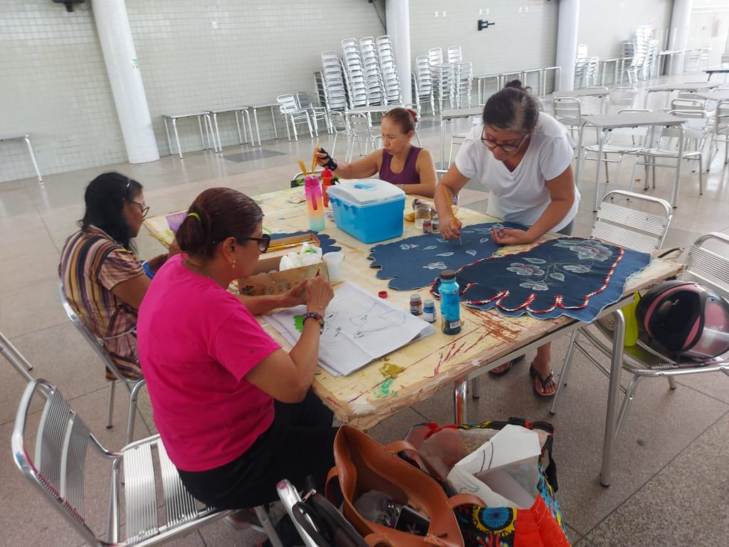 Grupo em oficina de artesanato, com foco em mãos trabalhando com tecidos e tintas em mesa coletiva