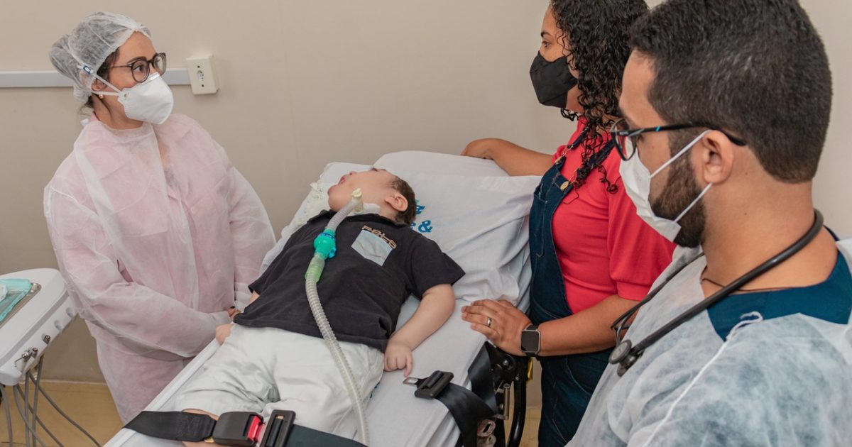 Equipe de profissionais da saúde em unidade básica, atendendo criança; profissionais representam fisioterapia, fonoaudiologia e odontologia, em ambiente de atenção primária