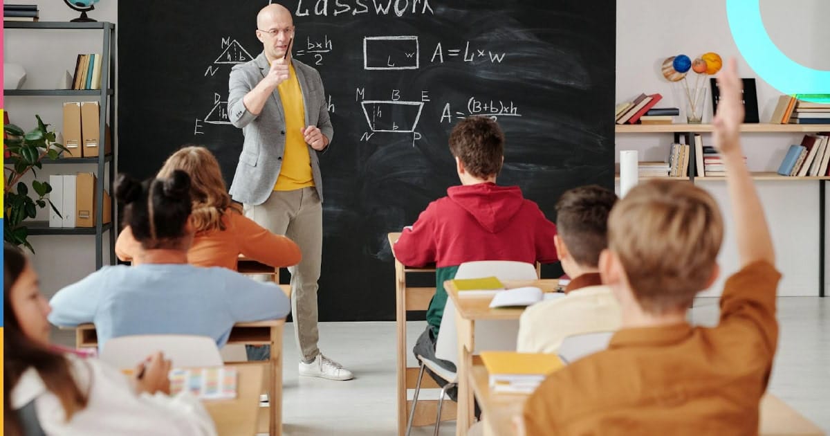 Professor em sala de aula interagindo com estudantes que levantam a mão.