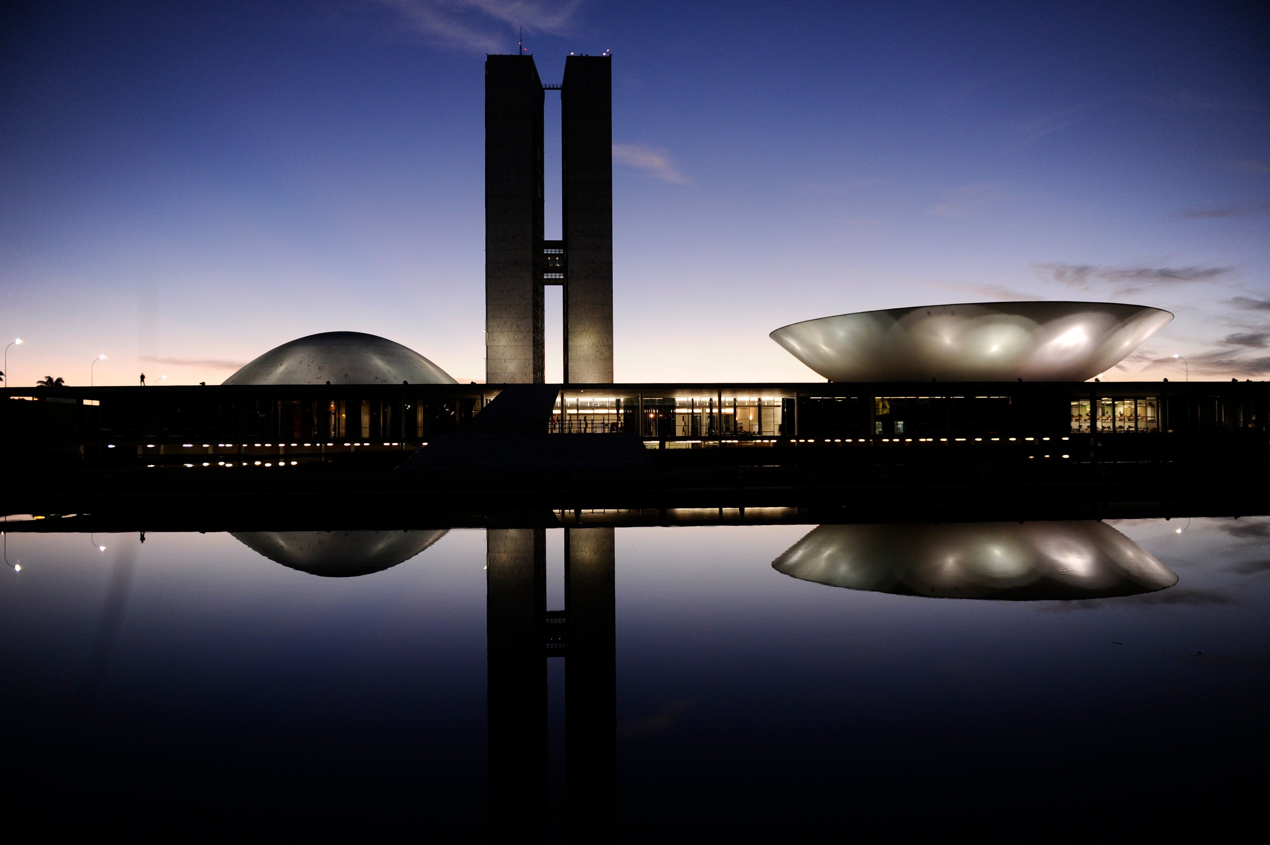 Vista aérea do Congresso Nacional em Brasília ao entardecer, com reflexo na água.