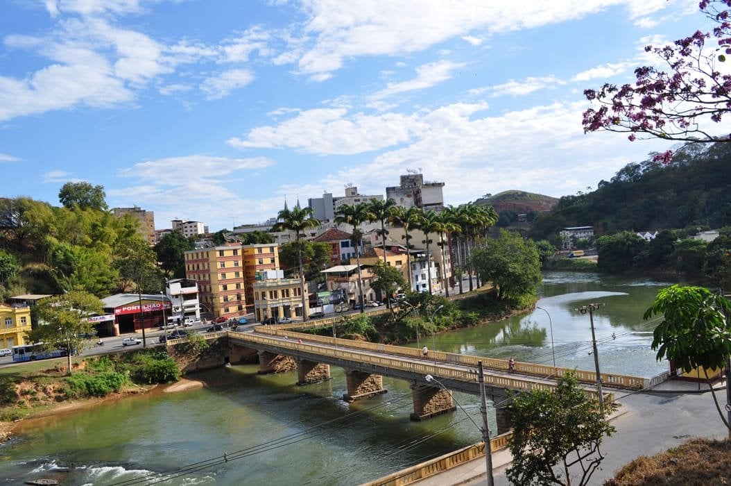 Vista aérea de Ponte Nova MG mostrando o centro urbano e a ponte sobre o Rio Piranga em dia ensolarado.