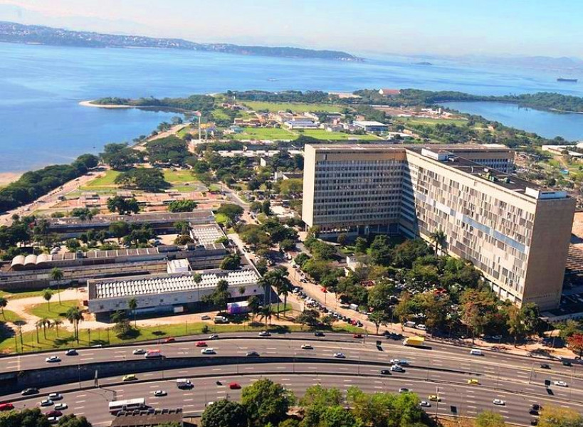 Vista aérea da Cidade Universitária da UFRJ na Ilha do Fundão, com edifícios, áreas verdes e água ao fundo