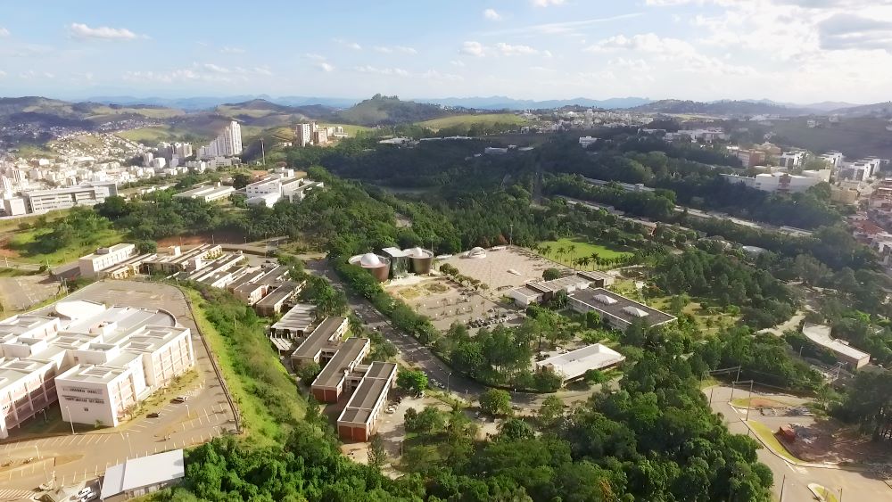 Vista aérea do campus da UFJF, com prédios e áreas verdes ao redor de morros, em dia parcialmente nublado