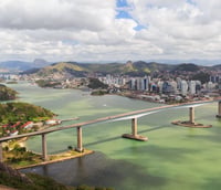 Vista aérea de Vitória ES ao pôr do sol, destaque para a Terceira Ponte e baía iluminada.
