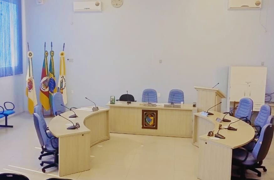 Interior de uma câmara municipal, com mesa diretiva e cadeiras em ambiente institucional