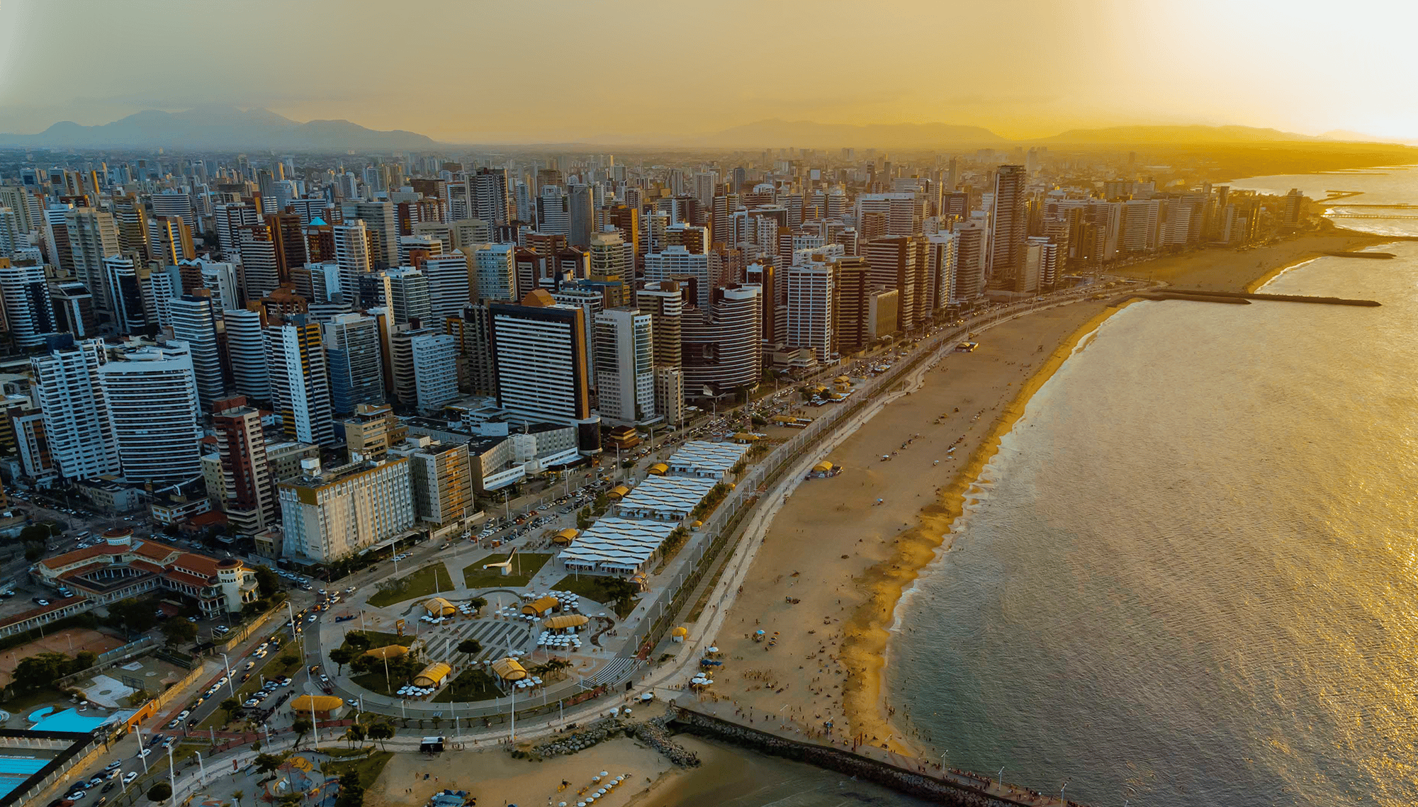 Vista aérea de Fortaleza ao entardecer, com prédios na orla da Avenida Beira-Mar.