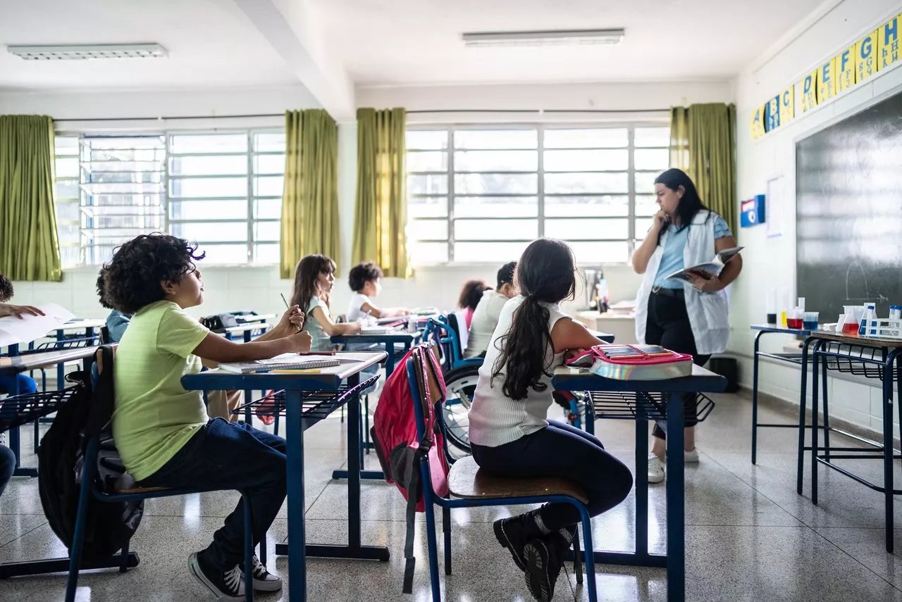 Sala de aula com estudantes e professor em atividade, luz natural entrando pela janela