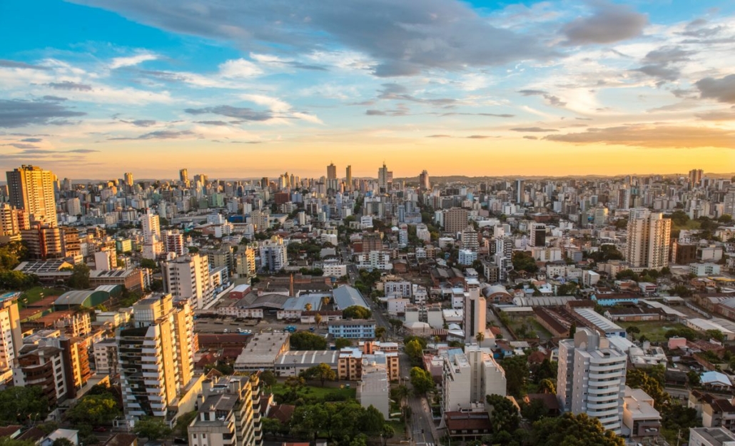 Vista aérea de Caxias do Sul com skyline urbano e colinas da Serra Gaúcha ao fundo, sob céu aberto